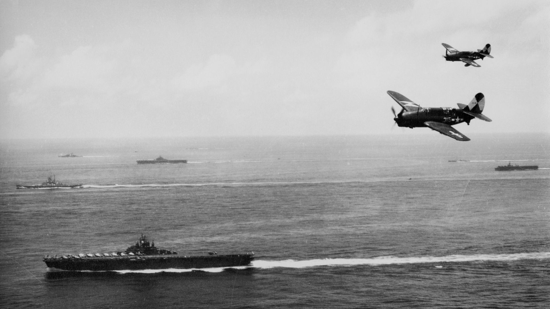 Two U.S. Navy Curtiss SB2C-4 Helldiver dive bombers of Bombing Squadron 83 (VB-83) fly against the backdrop of ships of Task Group 38.3 operating off Okinawa. VB-83 operated from USS Essex (CV-9), pictured in foreground, during the period March-September 1945. Note the geometric identification symbols (