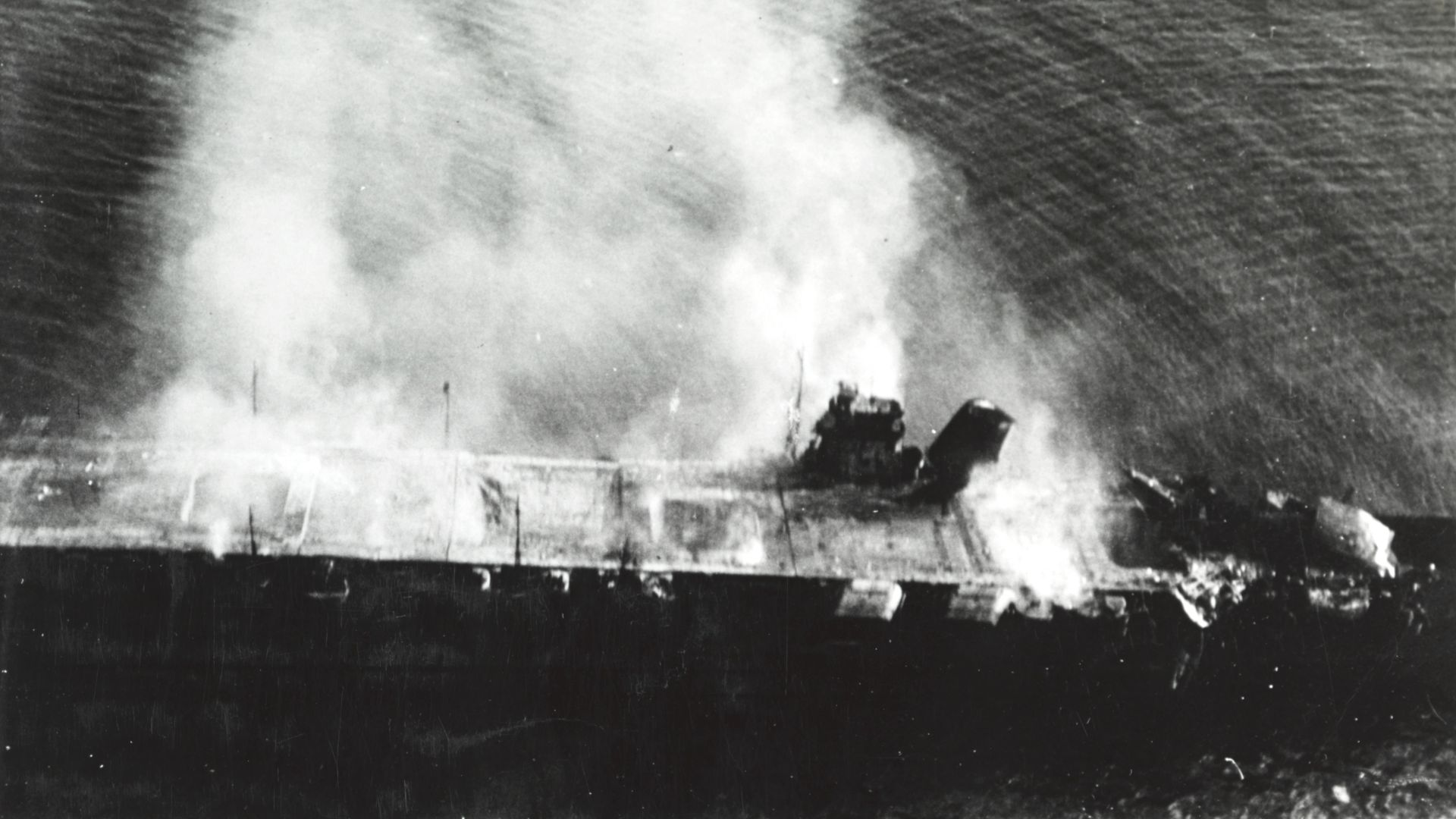 The burning Japanese aircraft carrier Hiryu, photographed by a Yokosuka B4Y aircraft from the carrier Hosho shortly after sunrise on 5 June 1942. Hiryu sank a few hours later. Note collapsed flight deck at right. Part of the forward elevator is standing upright just in front of the island, where it had been thrown by an explosion in the hangar.