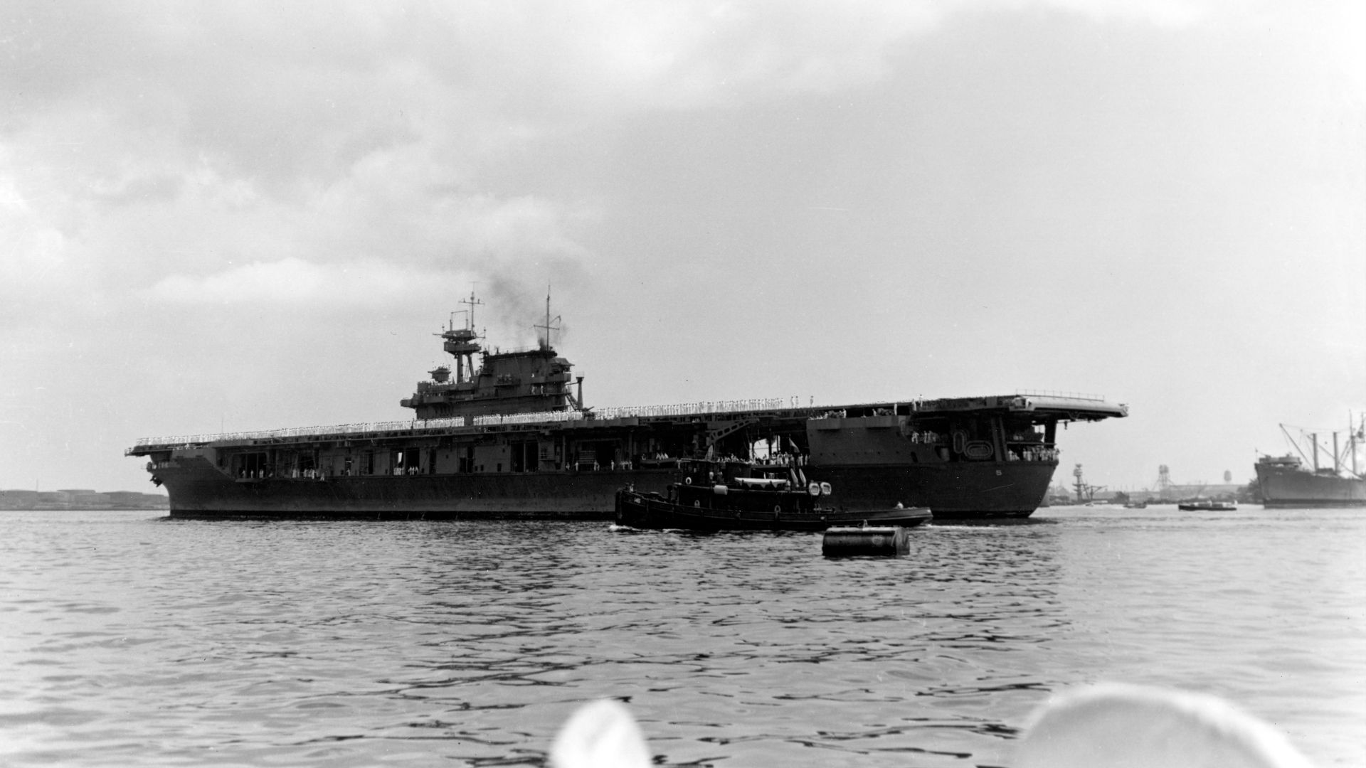 The U.S. Navy aircraft carrier USS Yorktown (CV-5) arrives at Pearl Harbor after the Battle of Coral Sea on 27 May 1942, with her crew paraded in whites on the flight deck. After hasty repairs, she departed on 30 May to take part in the Battle of Midway. The tug USS Hoga (YT-146) is in the center foreground. The mainmast of the sunken battleship USS Arizona (BB-39) is visible in the distance, just right of Yorktown´s stern. The carrier is wearing the standard Camouflage Measure 12.