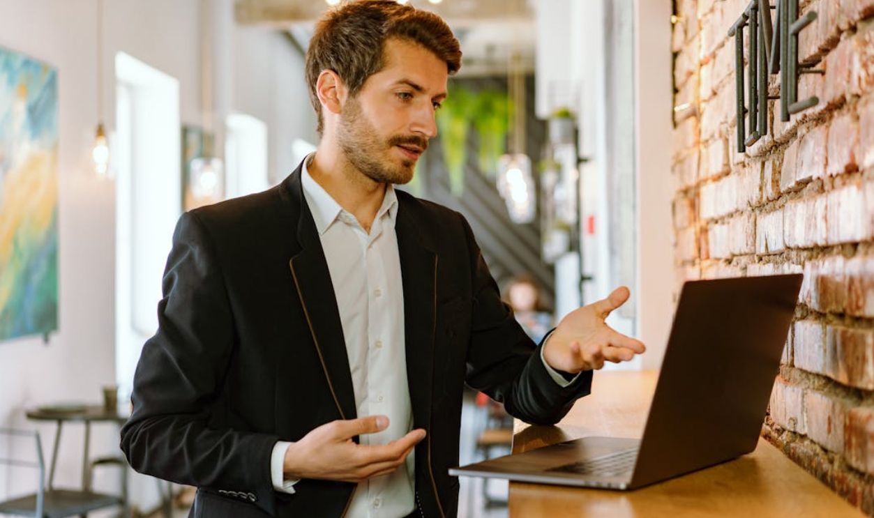 Young man in a black jacket engaged in an online meeting at a trendy cafe, gesturing while looking at the laptop