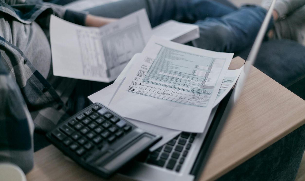 A young adult using a calculator and laptop to manage finances at home, surrounded by papers.