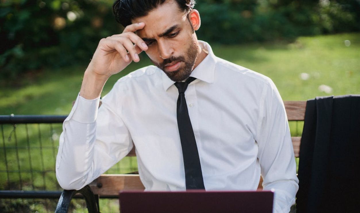 Bearded man in a white shirt and tie working on a laptop on a park bench