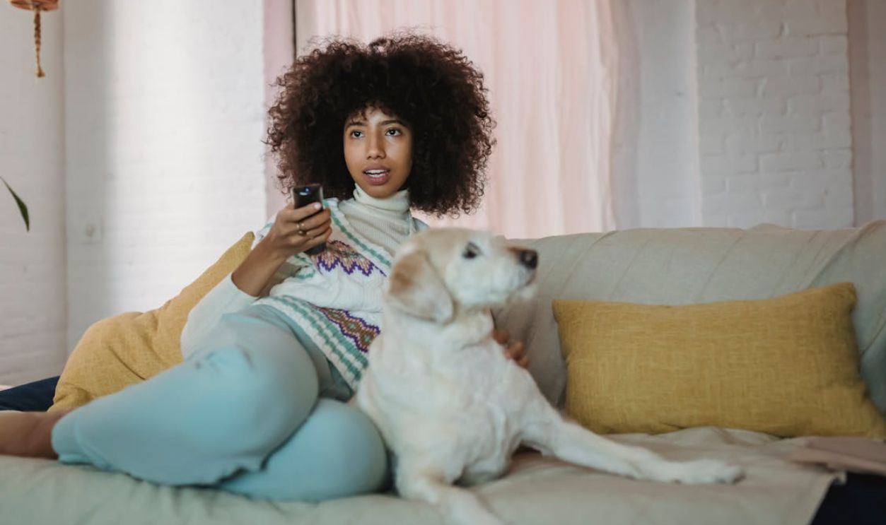 A young woman with curly hair enjoys a cozy moment with her dog on a living room couch.