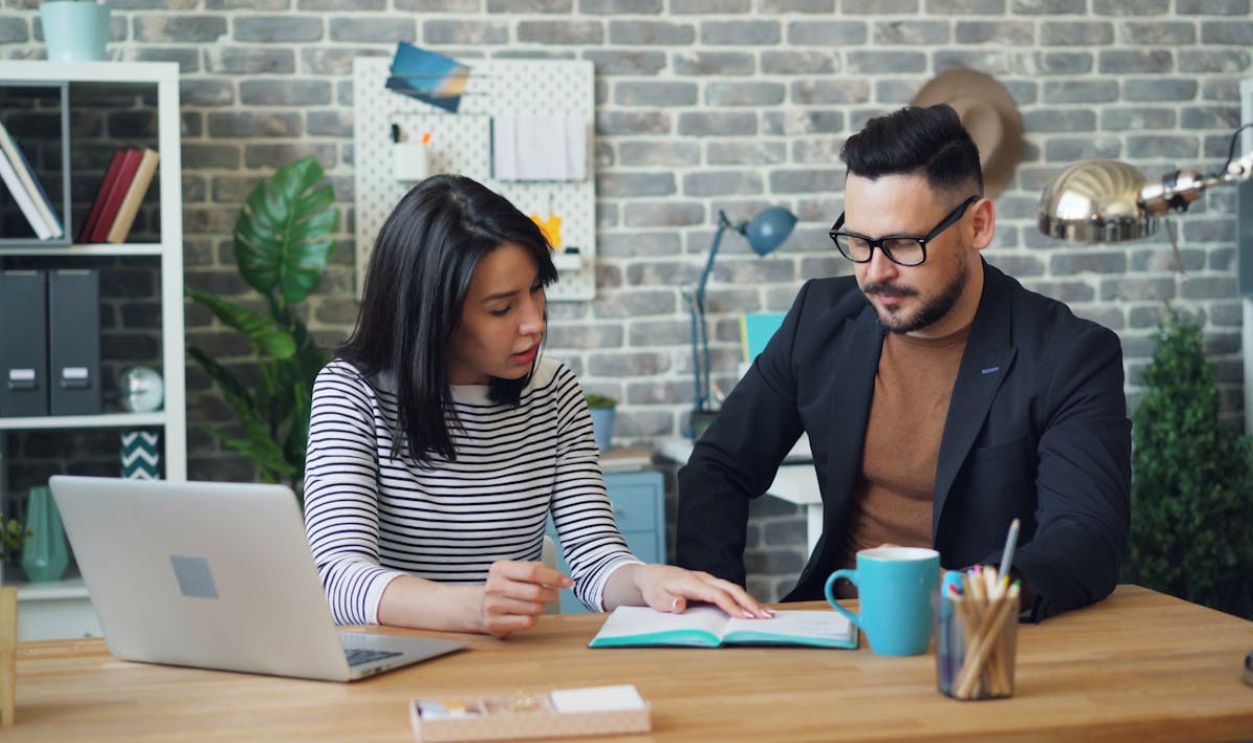 Two professionals discussing strategies at a modern office desk with a laptop.