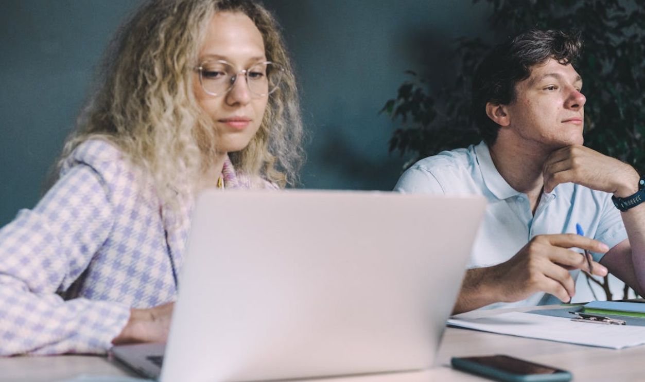 Two colleagues collaborate at a desk with laptops and papers, suggesting teamwork and productivity.