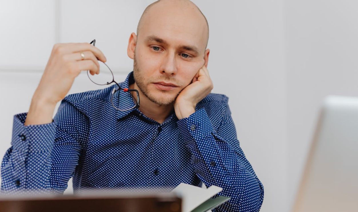 Bald businessman in blue shirt thinking at desk with glasses and laptop