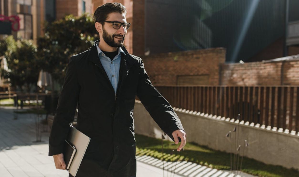 Businessman walking outdoors with a smile, holding a laptop, exuding confidence