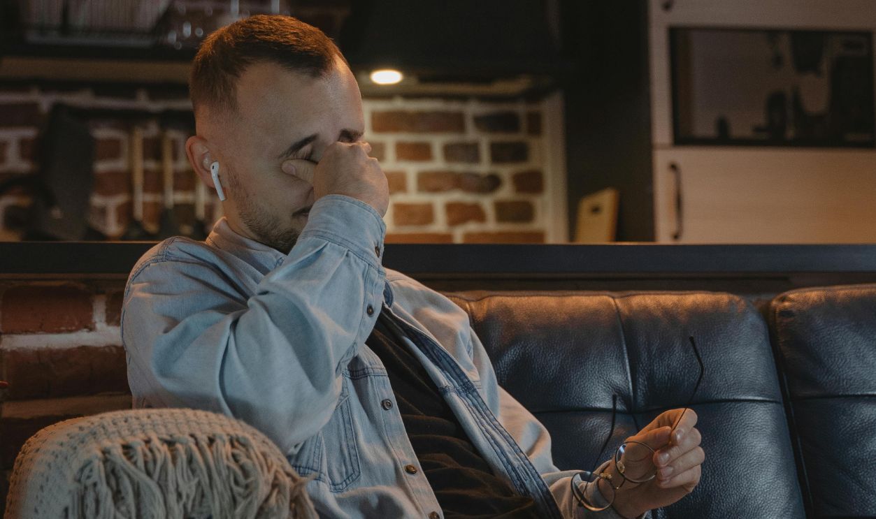 A bearded man in a denim jacket sits on a couch indoors, appearing tired while using a laptop