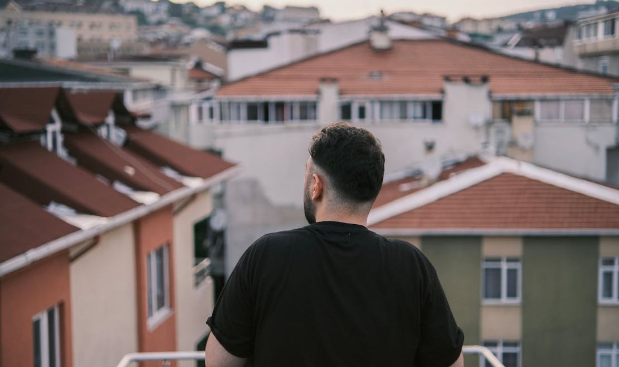 A man stands on a balcony gazing at a cityscape of residential rooftops during the day