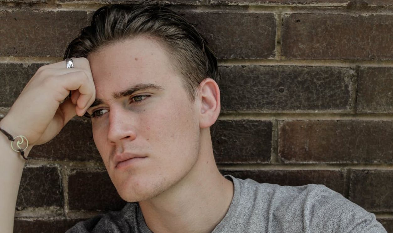Pensive young man leaning against a brick wall, deep in thought.