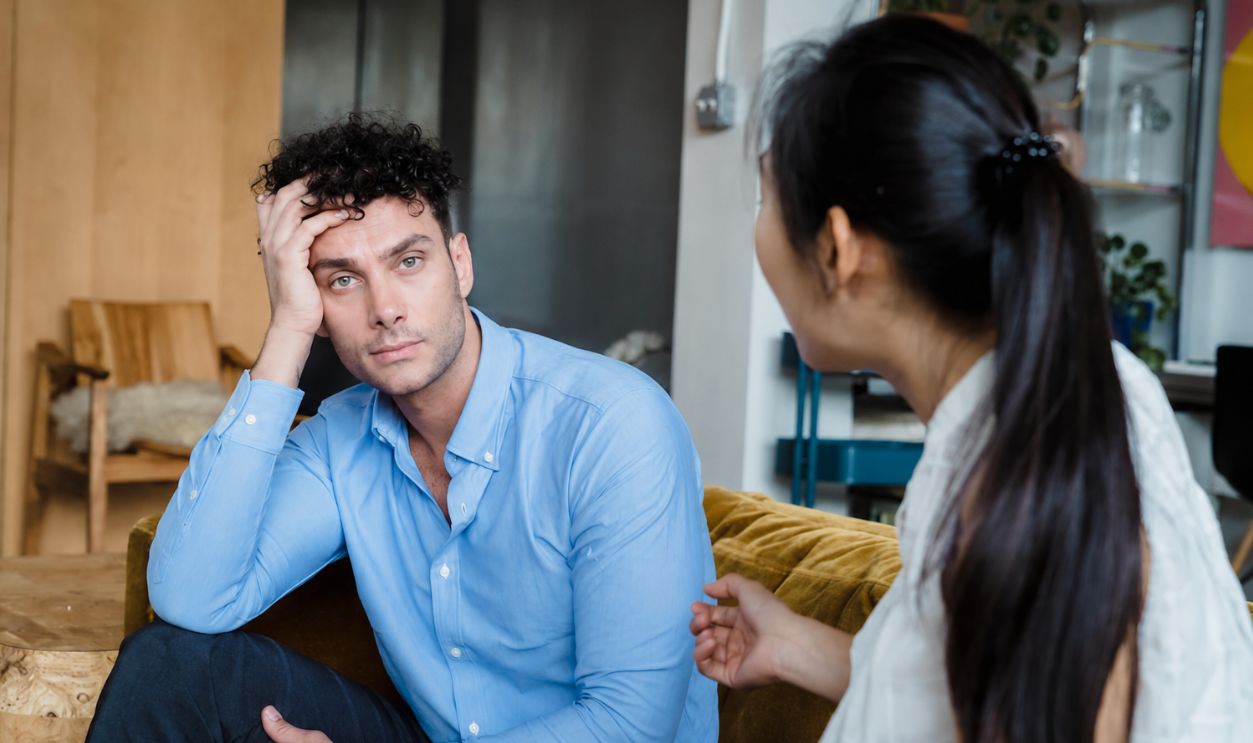 A tired man in a blue shirt listens while a woman talks indoors. Modern living room
