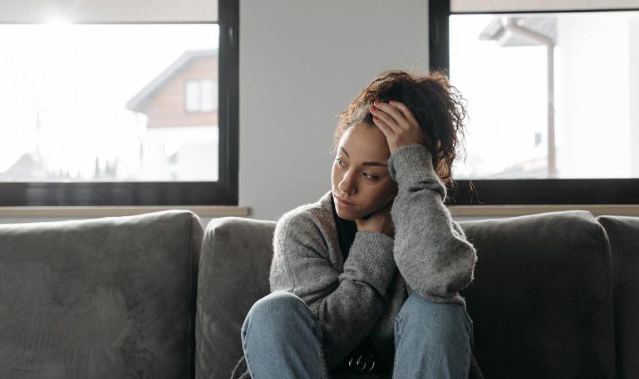 Thoughtful woman in cozy sweater sitting on sofa indoors by the window.