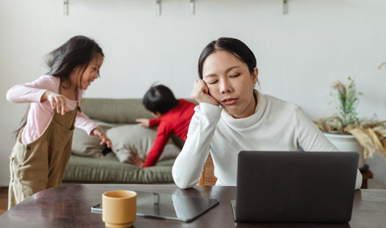A tired mother working remotely on her laptop while children play in the background.