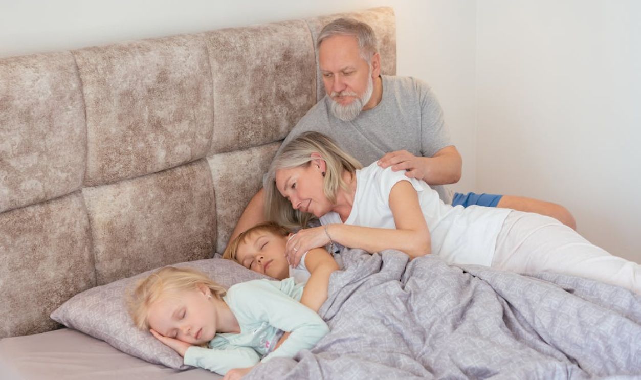 Grandparents lovingly watch over sleeping grandchildren on a cozy bed indoors.