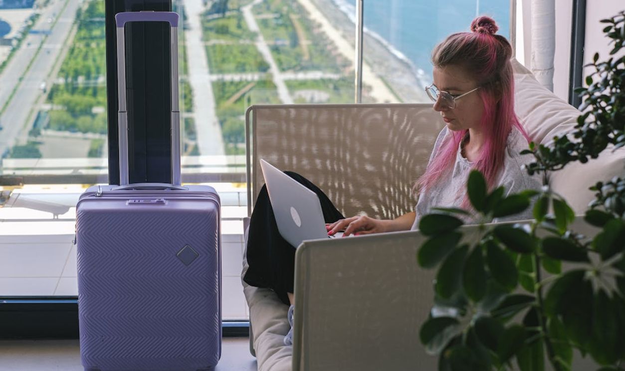 Woman with pink hair working on laptop in hotel room overlooking scenic landscape. Travel and freelance concept