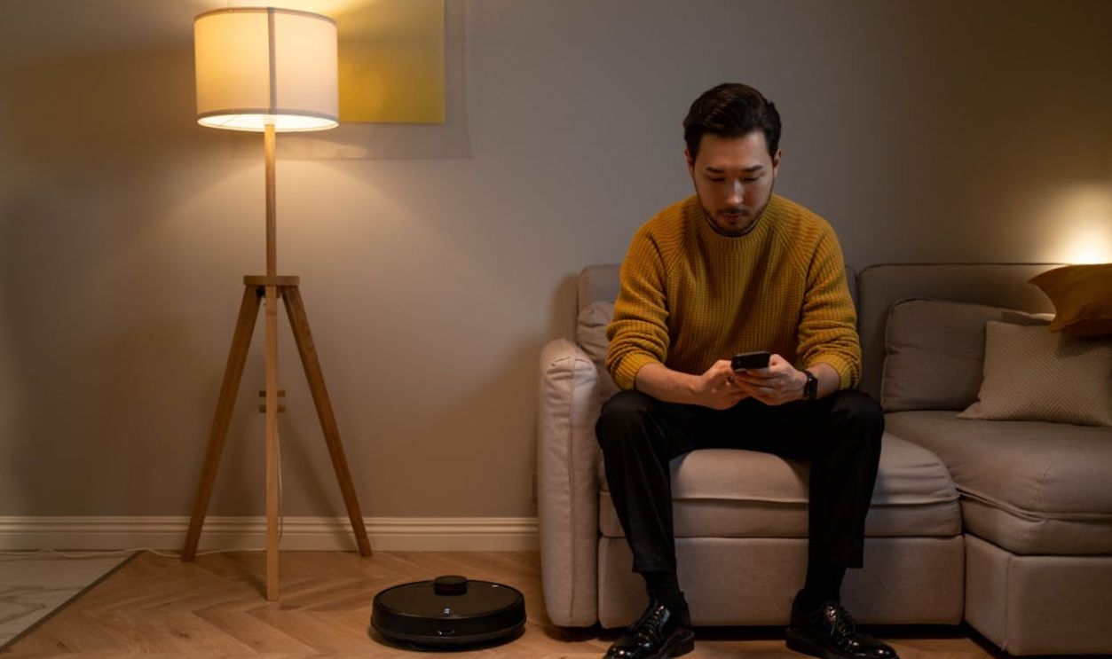 Asian man in a mustard sweater sits on a sofa, texting on his phone with a stylish lamp illuminating the space
