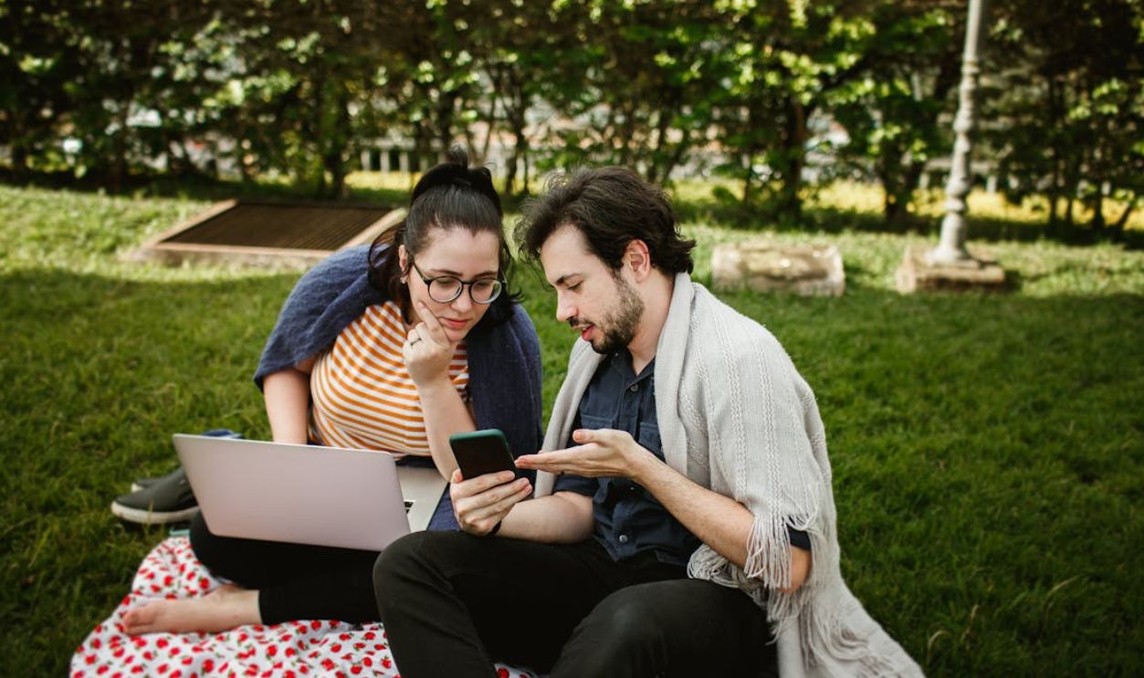 Young couple sitting outdoors, using laptop and smartphone on a sunny day in the park