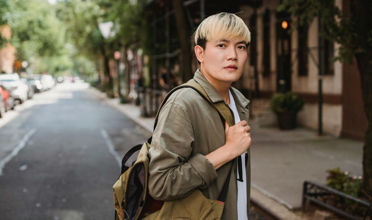 A stylish man with dyed hair and backpack walking down a city street on a sunny day.