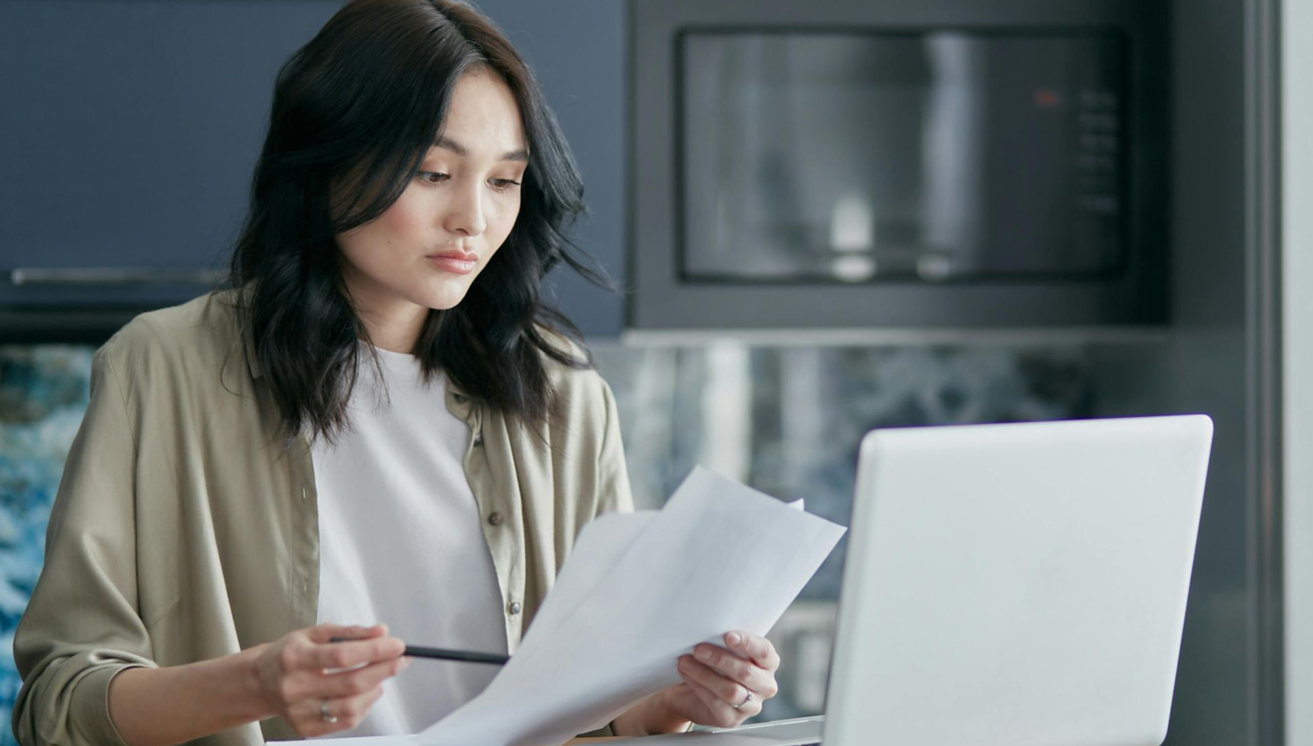 Concentrated woman reviewing documents and working on a laptop at home office setup in kitchen.