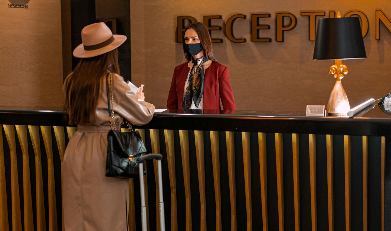 A woman speaks to a receptionist wearing a mask at a hotel reception