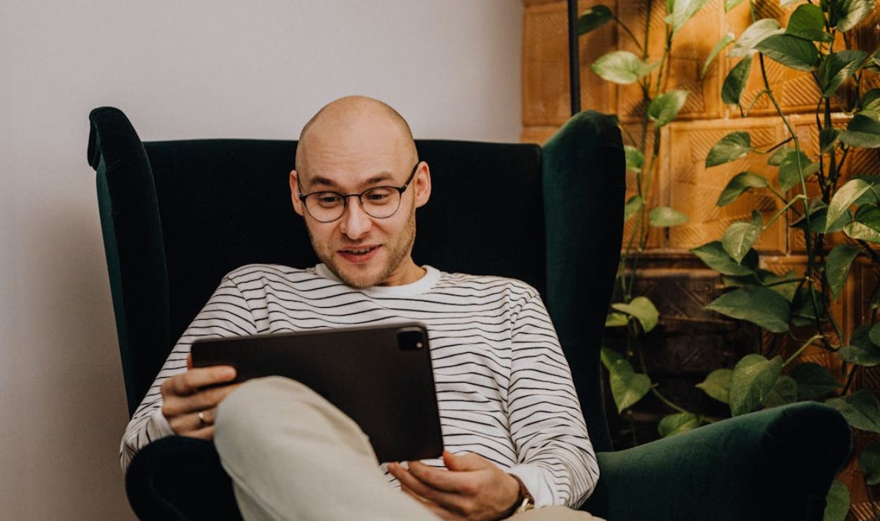 Bald man with eyeglasses sits comfortably in a green armchair indoors, using a tablet