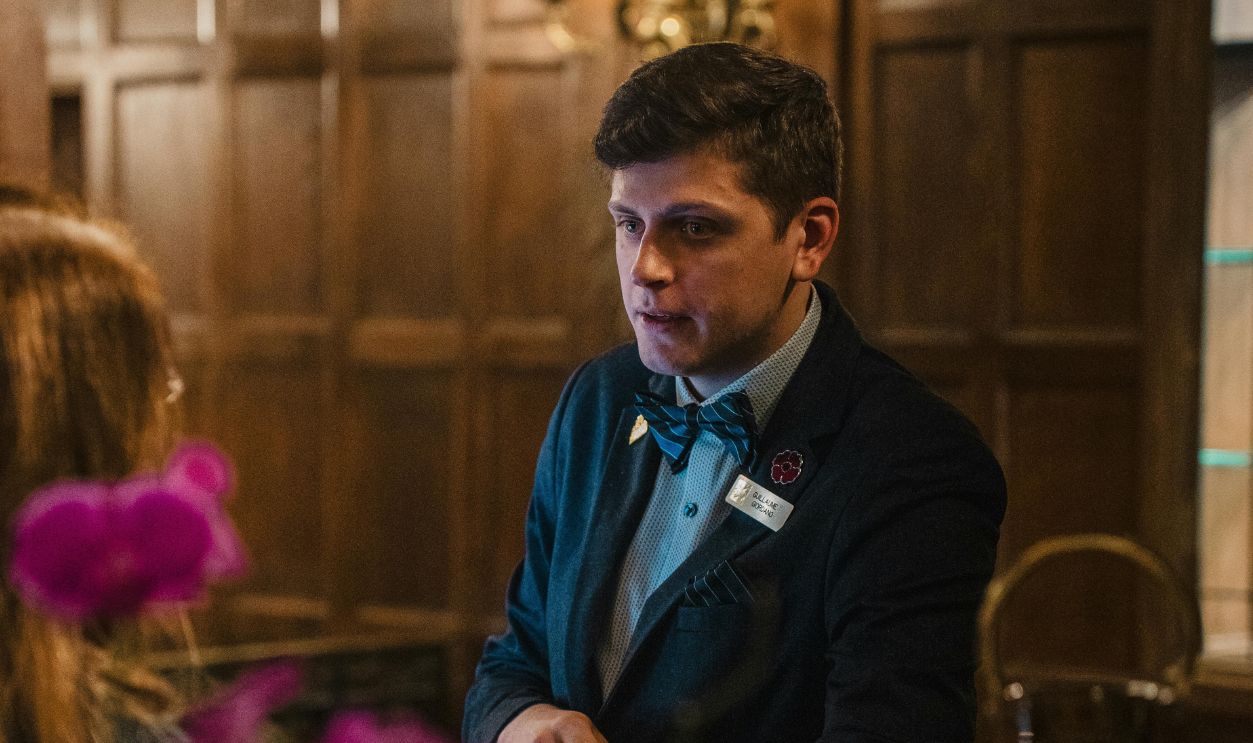 Polite hotel receptionist in formal attire at a warmly lit wooden reception desk