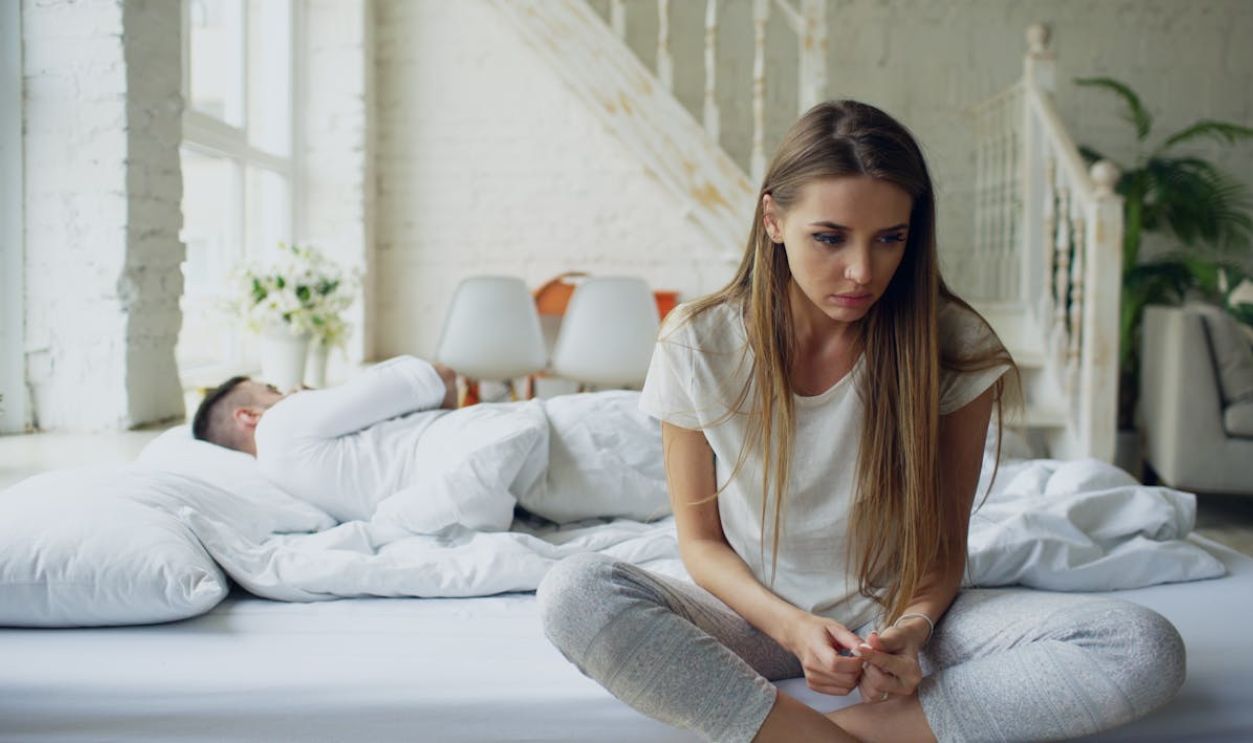 Woman sitting on bed looking thoughtful while man sleeps, symbolizing relationship tension.