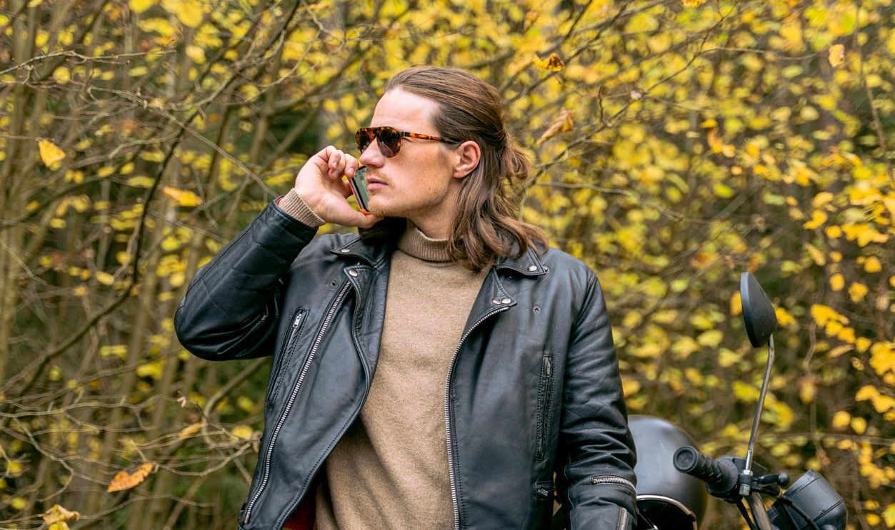 Man in leather jacket making a phone call beside a motorcycle in a fall forest setting