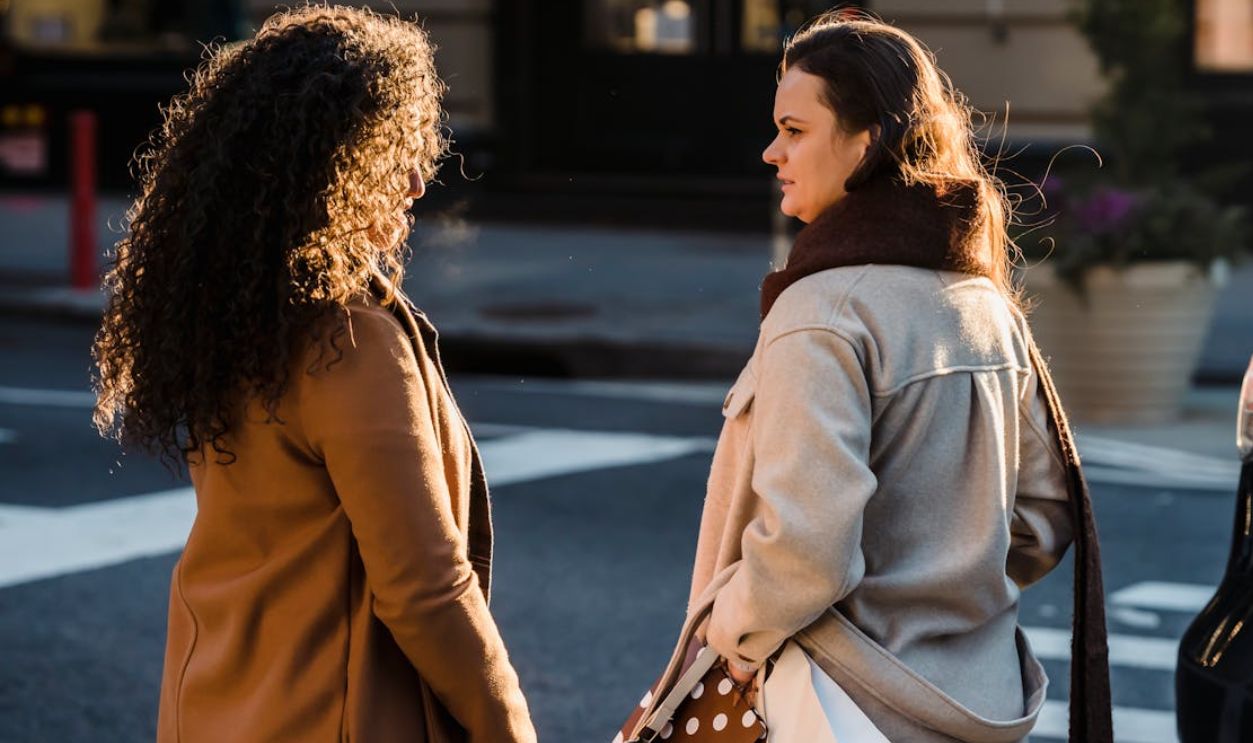Two women engage in conversation on a sunny urban street, showcasing friendship and casual fashion