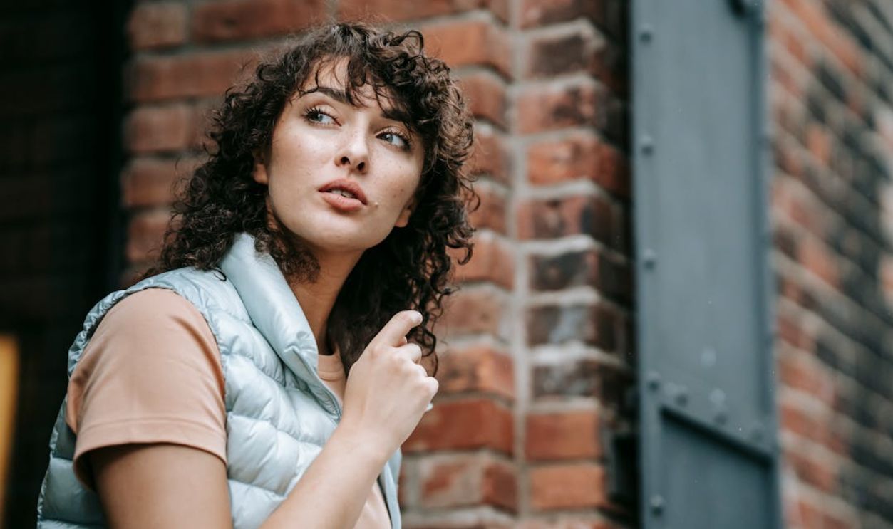 Curly-haired woman reading outdoors with a thoughtful expression against a brick wall
