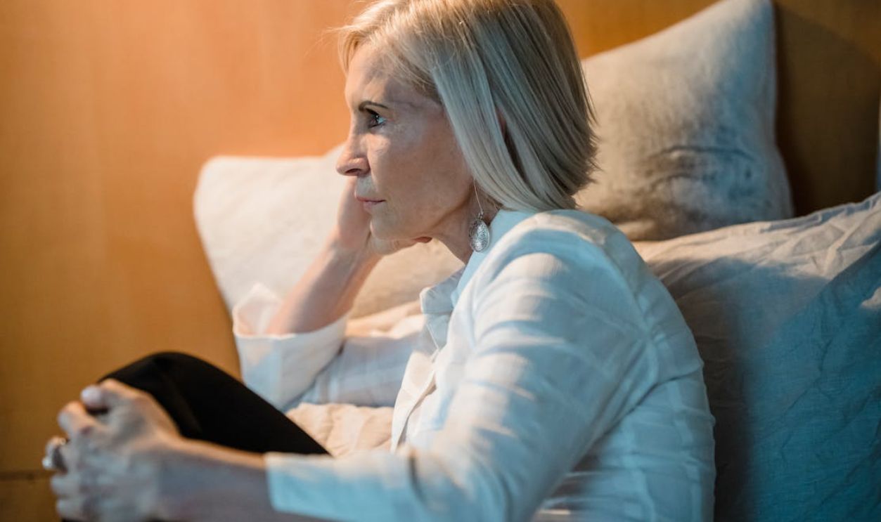 Elderly woman in deep thought, sitting on a bed with soft pillows, wearing white shirt