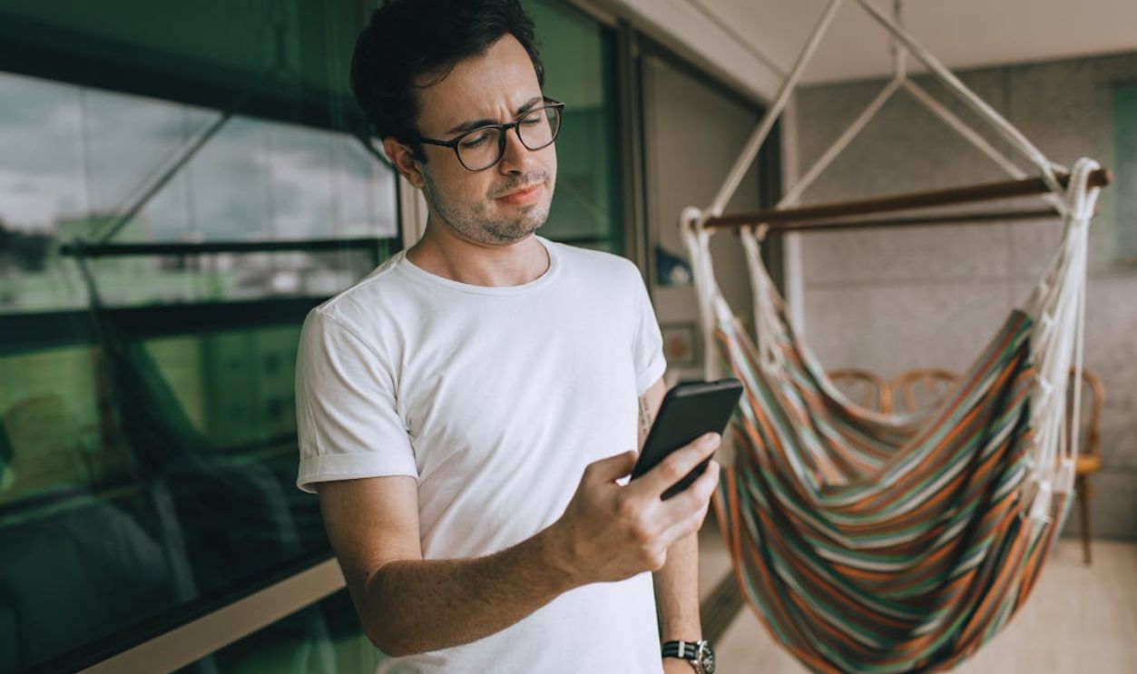 Young man with eyeglasses checking phone on balcony by a hammock