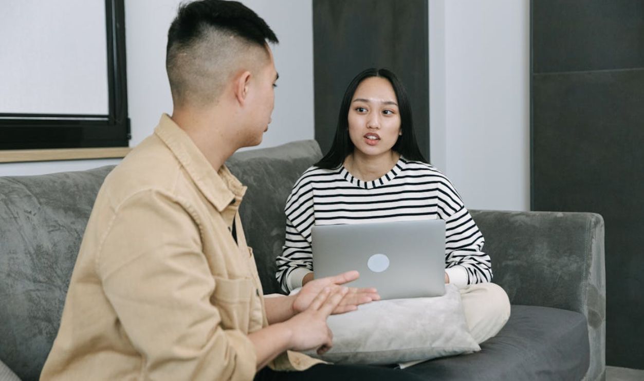 Two young adults engaged in a conversation while sitting on a sofa with a laptop