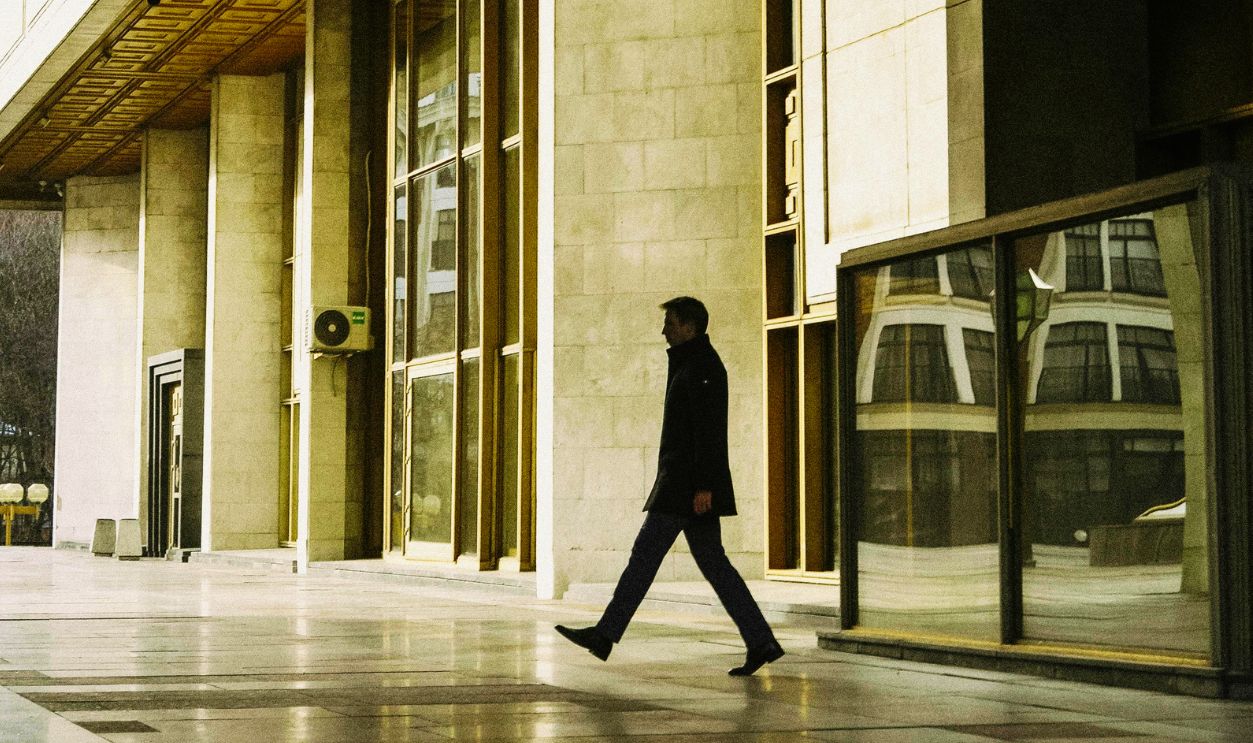 A man in a coat walks through modern architecture in an urban setting with reflective surfaces