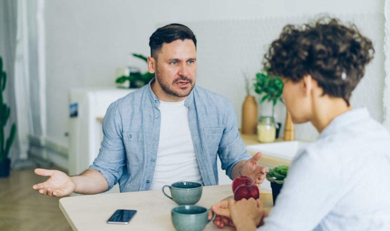 Two adults engage in a conversation at a kitchen table with coffee cups