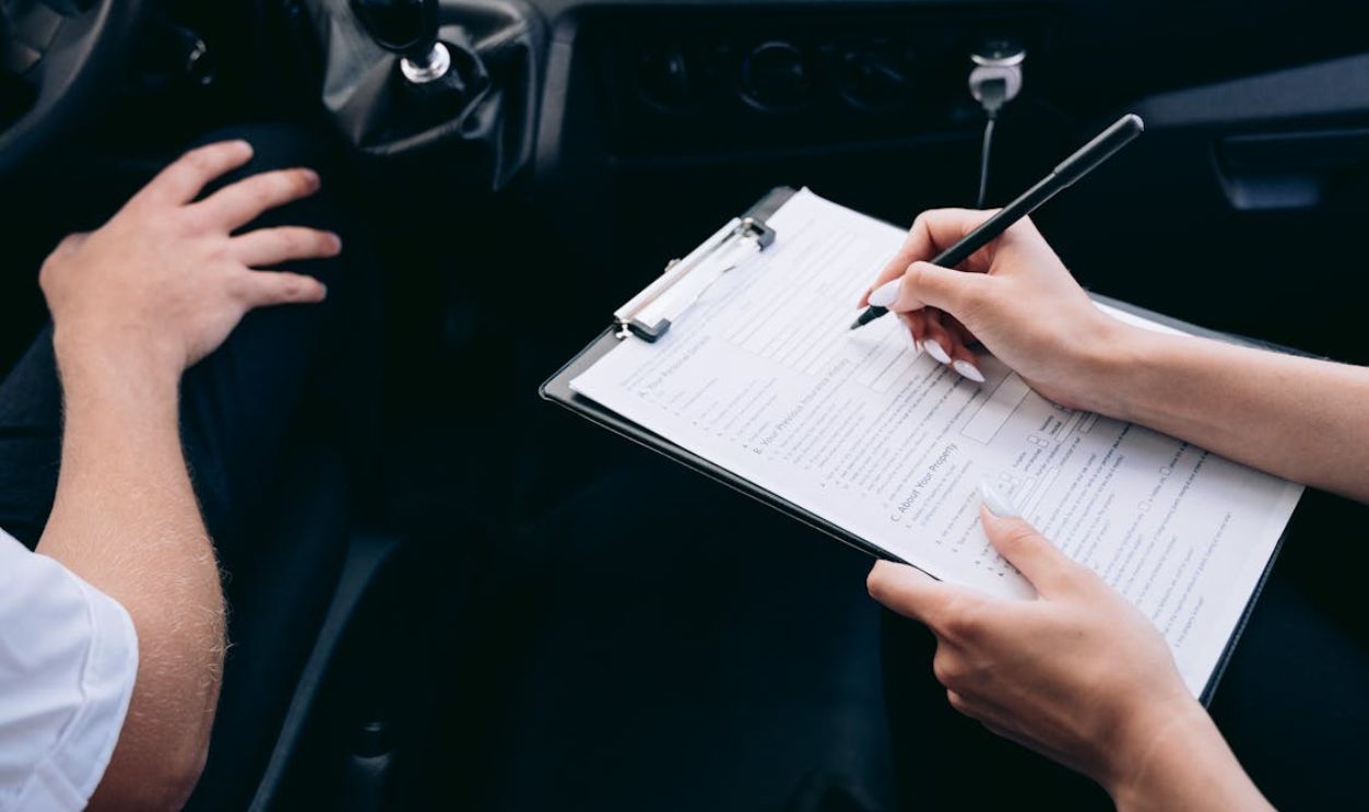 Close-up of a person writing on a clipboard inside a car, showing hands and a gear shift.