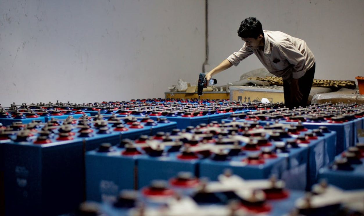 A worker checking many industrial batteries inside a facility. Indoor, industrial setting.