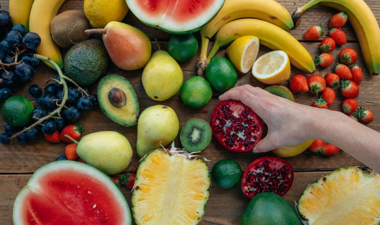 Colorful assortment of fresh fruits on wooden surface with a hand holding a pomegranate half