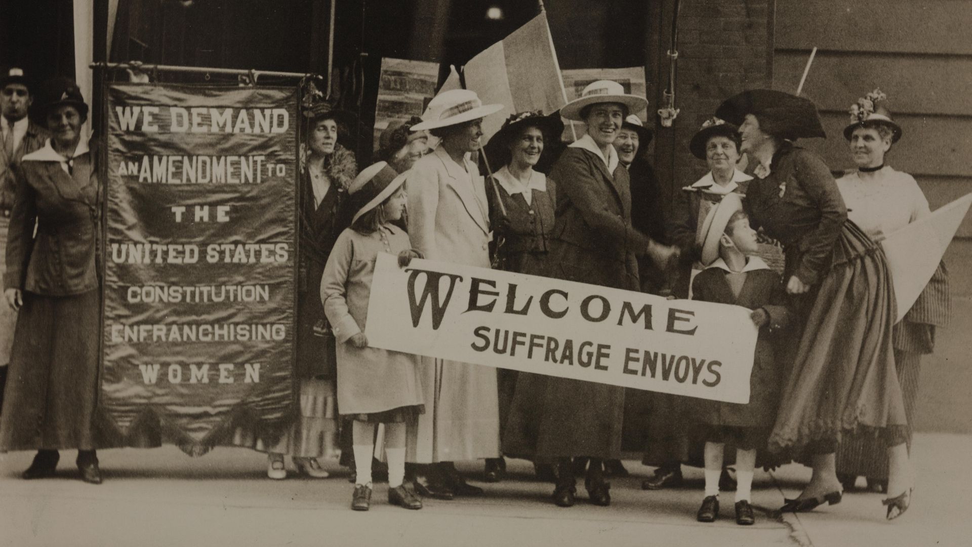 Summary: Photograph of group of smiling women and two children standing with banners on sidewalk. Banner on left: