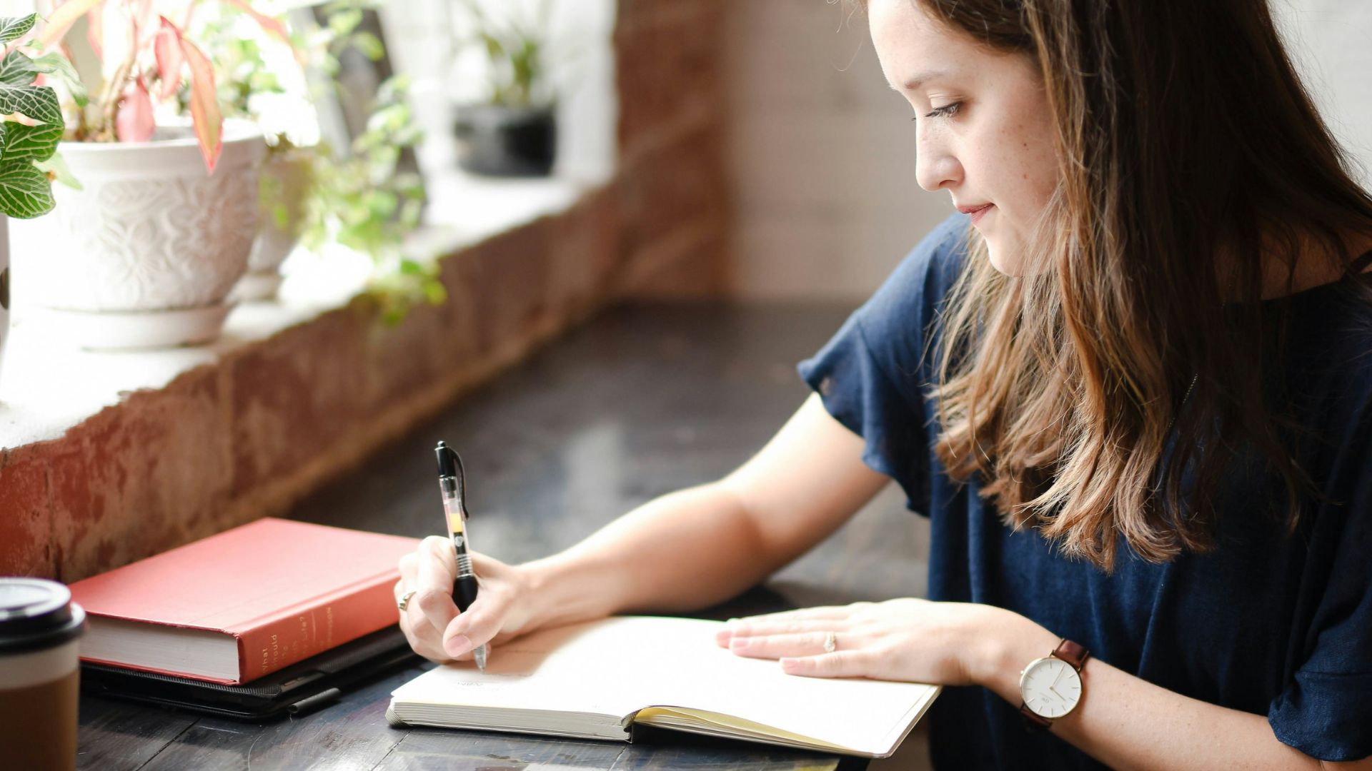 woman sitting in front of black table writing on white book near window