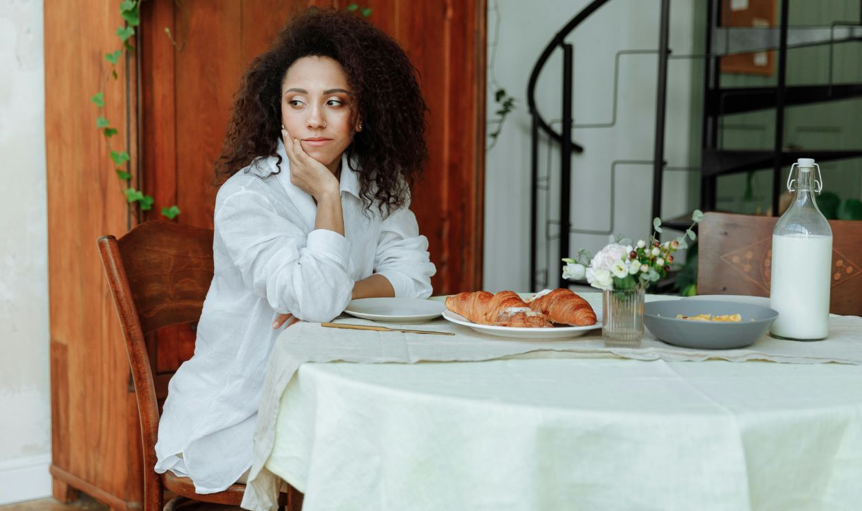 A woman with curly hair sits at a breakfast table with croissants and milk