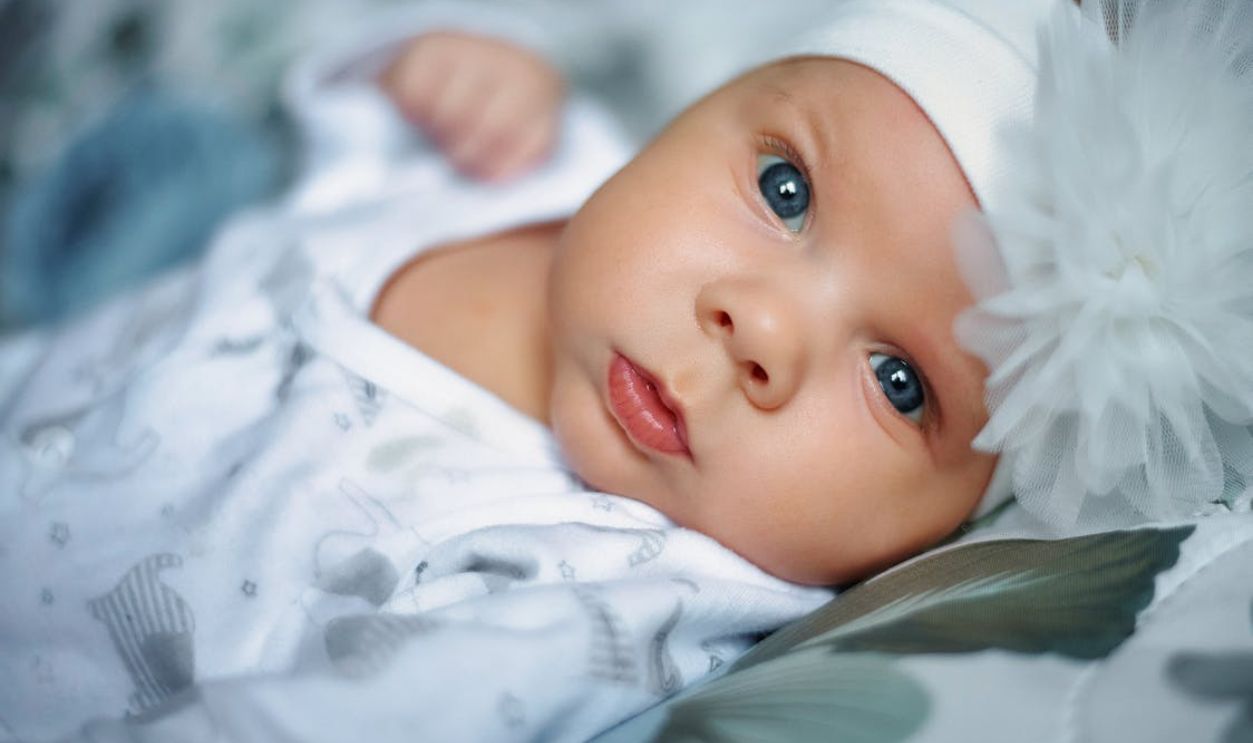 Cute newborn baby with blue eyes lying on bed, wearing white headband and flower.
