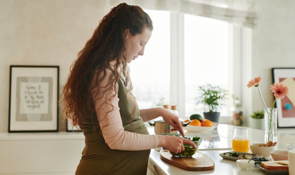 Pregnant woman with curly hair prepares a meal on kitchen counter, surrounded by fresh ingredients.