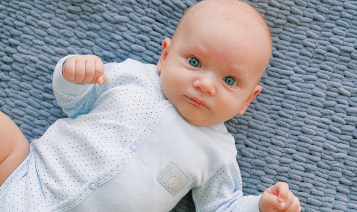 Cute baby in polka dot onesie lying on a textured blue blanket, looking up with a calm expression.