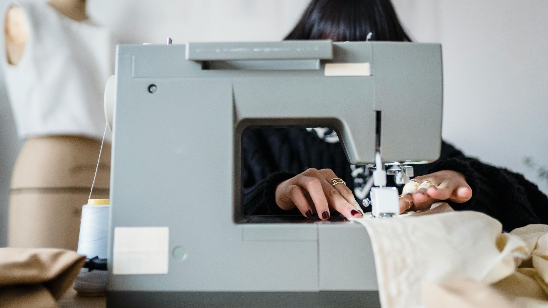 Unrecognizable female dressmaker with dark hair creating new fashion collection using sewing machine in light workshop