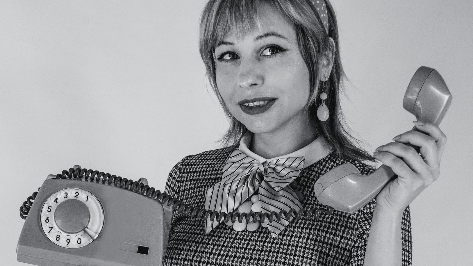 Caucasian woman in vintage attire holding a rotary phone, studio black and white.