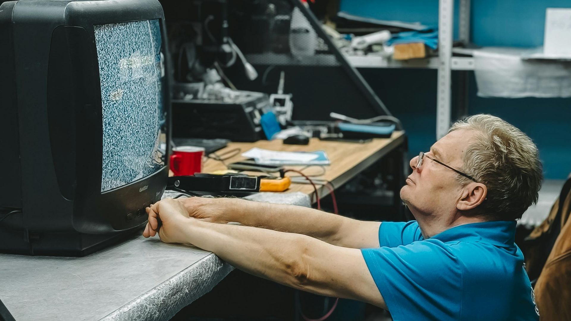 Man repairing an old television with static in a cluttered workshop setting.