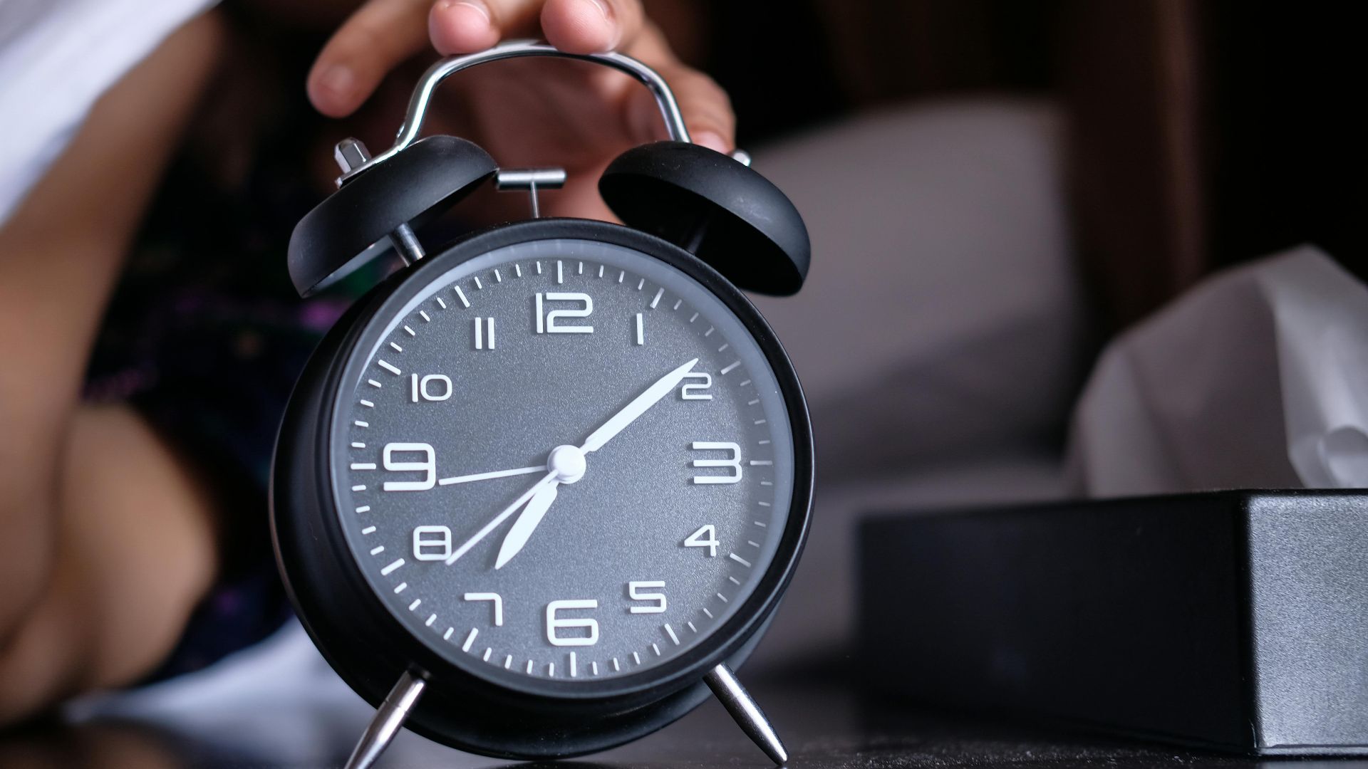A hand reaches out to turn off an analogue alarm clock in the early morning.