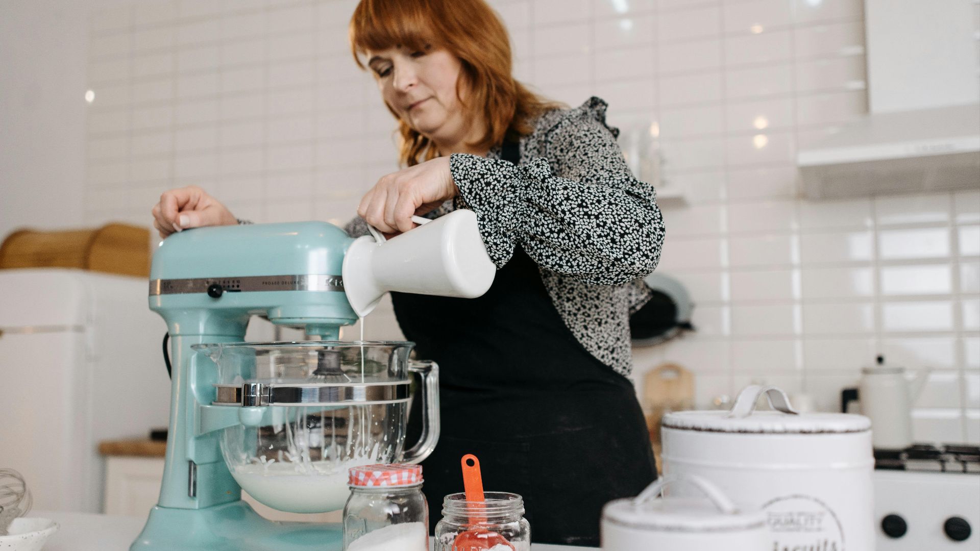 Woman pours ingredient into mixer bowl during food preparation in a modern kitchen setting.