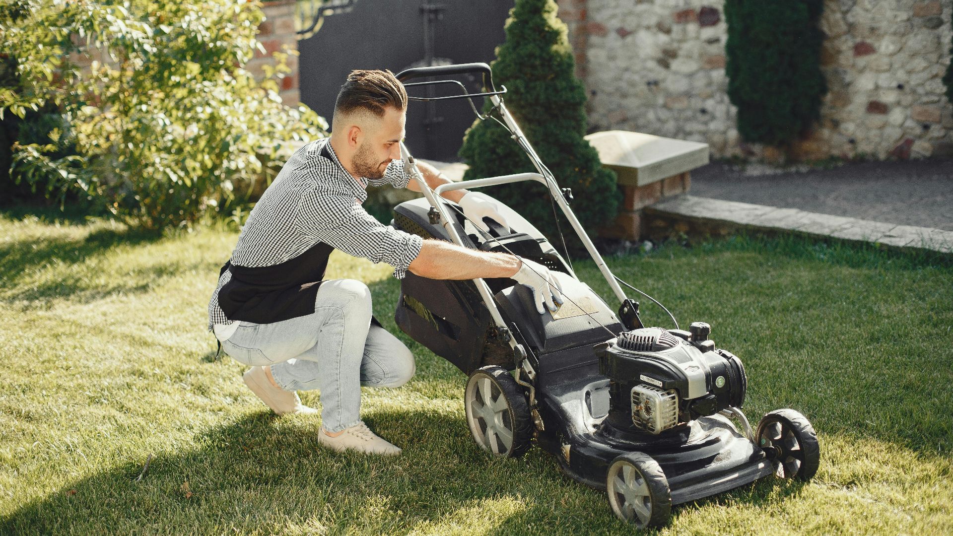 Man in summer attire using a lawnmower on a sunny day, showcasing gardening tools.